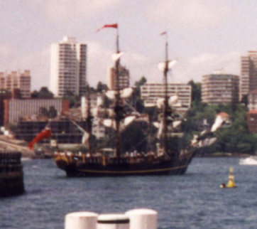 A ship with furled sails in a body of water with a city in the background