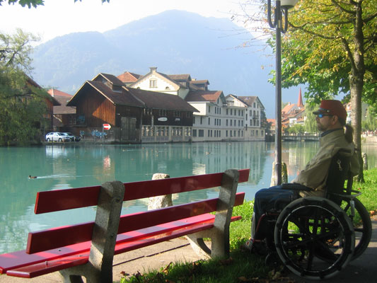 Gene sitting in his wheel chair next to a bench and a beautiful clear lake