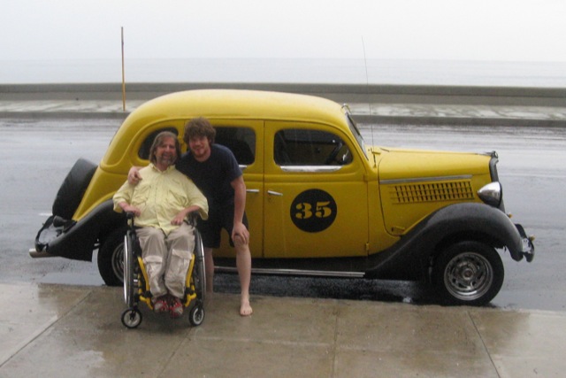 Gene and Pedro smile outside of Pedro's car, a yellow vintage style car with a black circle and the number 35 on it.