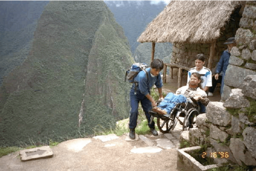 Alex and Jaime carry Gene in his chair along a path in Machu Pichu. Large mountains are seen in the distance.