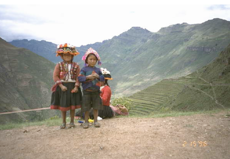 Young children native of Peru stand on a trail with mountains in the background. The children wear traditional clothing.
