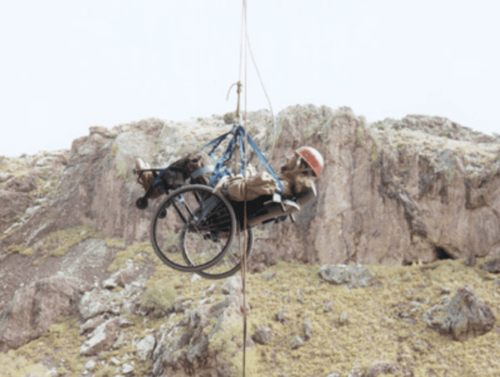 Gene in midair on the pully system that helps him ascend the cliff face. Gene is in his wheel chair while being lifted up.
