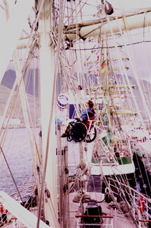 Gene being lowered with his wheelchair on a ship using a block and tackle system