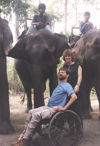 A woman holding on to a man in a wheel chair with a background of a group of people riding on the backs of elephant