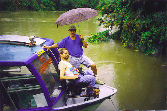 Two men one with an umbrella and the other sitting in a wheelchair n front of a boat on a river