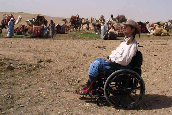 Gene relaxing in his wheel chair with Bedouin persons seen in the background on a desert plain