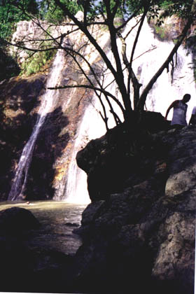 A waterfall in Thailand with a silhouette of a person standing near a tree in front of the falls