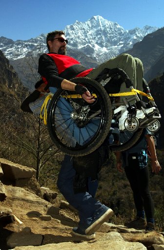 A man carries another man in a wheel chair over his shoulders while climbing a mountain.