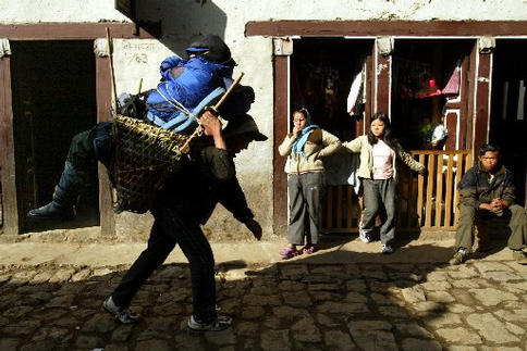 A sherpa carries a man in a bamboo basket through a town street. Towns people look watch curiously.