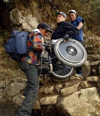 A man in a wheel chair is being carried of a rocky terrain by two sherpas.