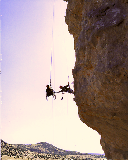 silhouette of Gene ascending a cliff face in his wheelchair with the assistance of a guide